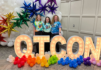 Staff in rainbow attire posing by light up letters that read QT-Con.