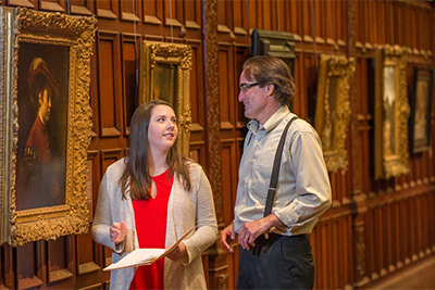 A woman and a man converse while observing ornate framed paintings in an art gallery.