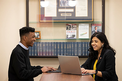 Two college students engaged in a lively discussion while working on laptops at a table, with bookshelves and framed documents in the background.