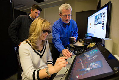 A woman wearing 3D glasses interacts with a touchscreen display while a man provides guidance, and another man watches in the background.