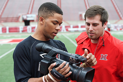 Two young men discussing video footage on a camera at a sports field.