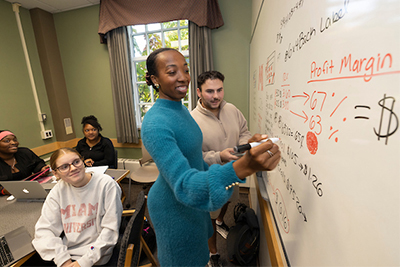 A student in a blue sweater actively writes on a whiteboard during a classroom discussion, while classmates observe and take notes.