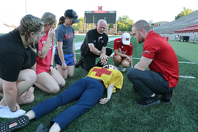 A group of students and instructors participate in a first aid training session on a football field, demonstrating proper techniques on a lying figure.