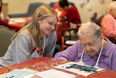 A young volunteer engages with an elderly woman as they admire a colorful artwork during a creative activity session.