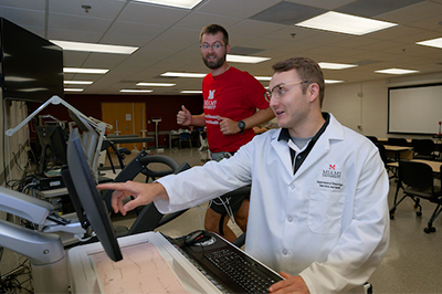 A researcher in a lab coat points at a monitor while a participant in a red shirt runs on a treadmill in the background.