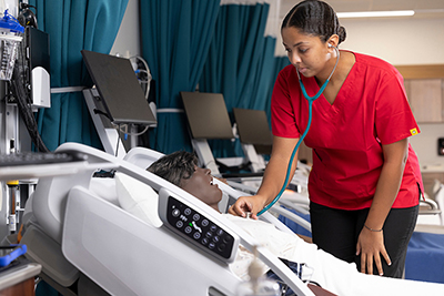 Nursing student in red scrubs monitoring a patient mannequin in a healthcare simulation lab.