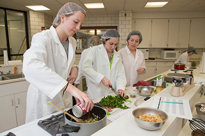 Students in white lab coats prepare a meal in a cooking lab, showcasing teamwork and culinary skills.