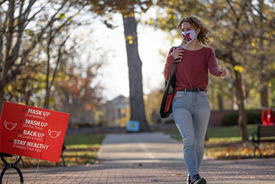 A student wearing a mask walks on campus next to a sign promoting COVID-19 safety guidelines.