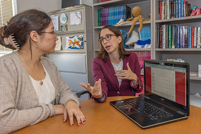 Two women engage in a conversation at a desk, with a laptop open in front of them, surrounded by shelves of books and educational materials.