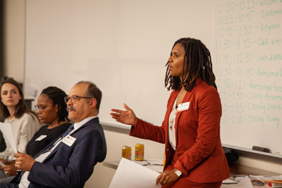 A woman in a red suit speaks confidently at a panel discussion, while two attendees listen attentively. A whiteboard with agenda items is visible in the background.