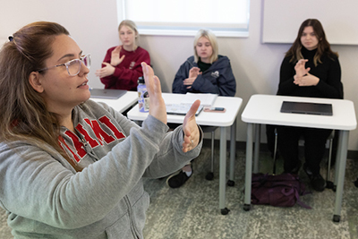 A student gestures in front of a classroom while peers observe, participating in a sign language lesson.