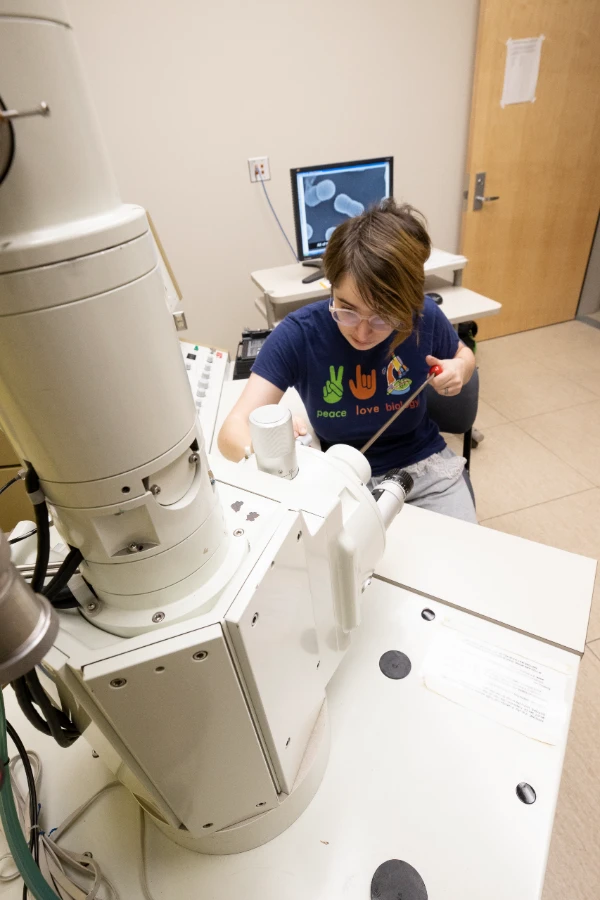 Person wearing a purple shirt, sitting in front of a machine in a lab.