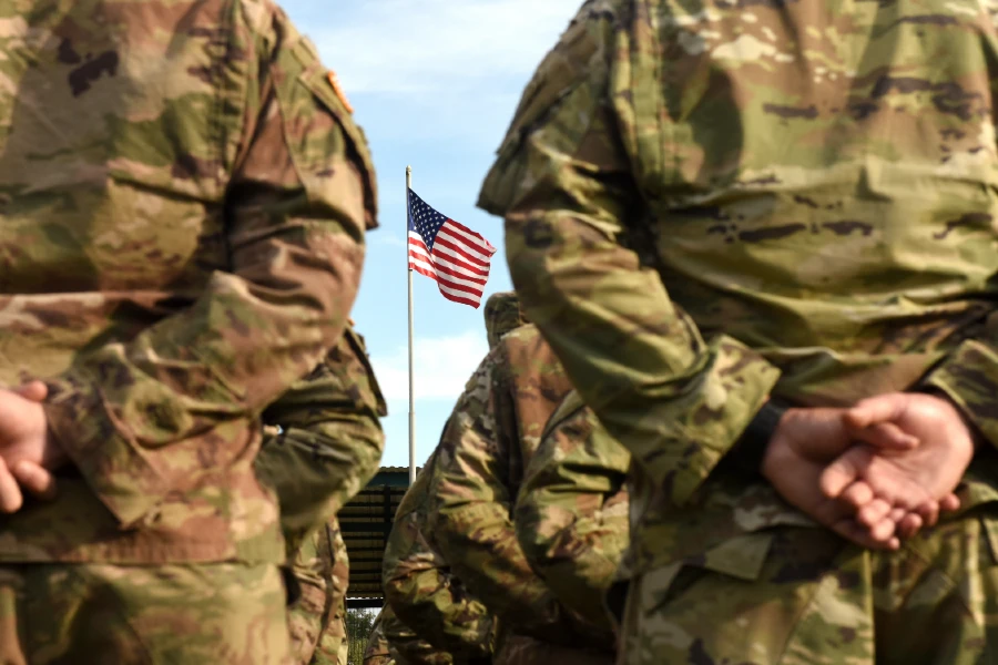 Soldier wearing fatigues facing the American flag, with their hands clasped behind their backs.