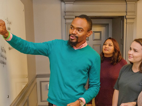 Male professor pointing at a white board instructing two female students.