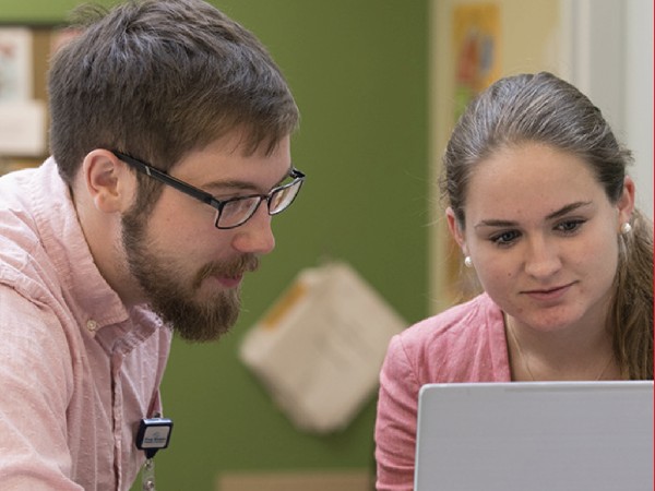 Male professor engaging with a female student who is working at a computer.