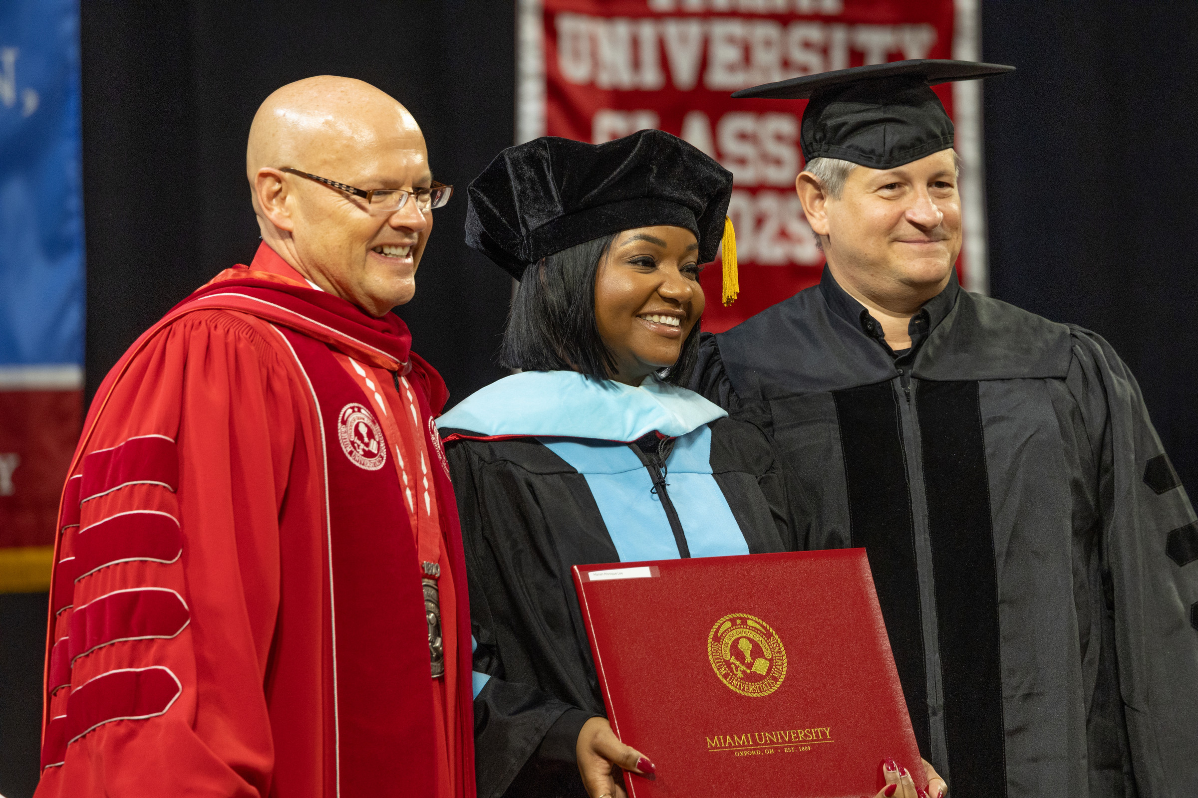 A doctoral graduate smiles while holding a diploma at a graduation ceremony.