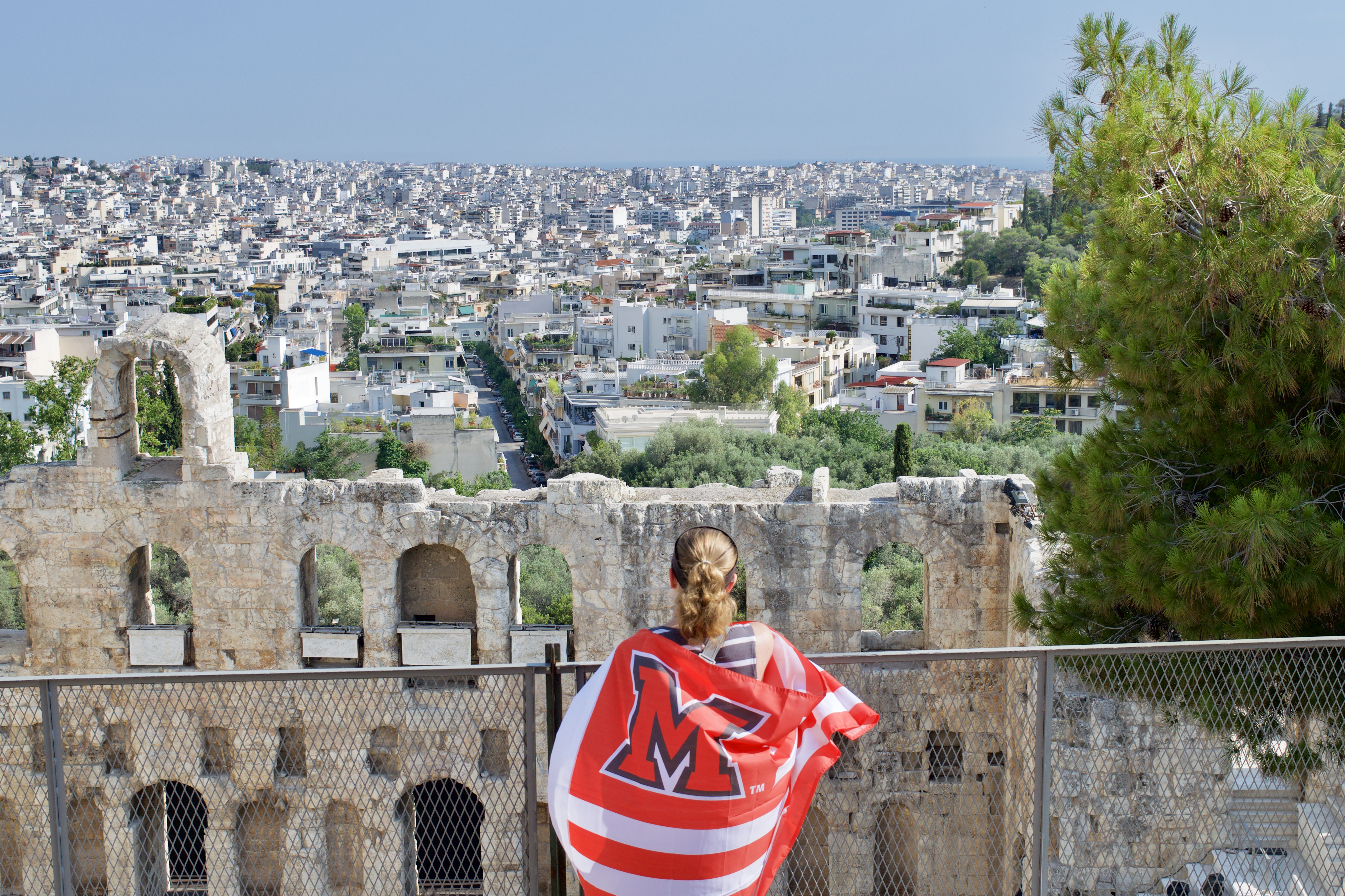 A student draped in a Miami flag looks out over a city in Greece.