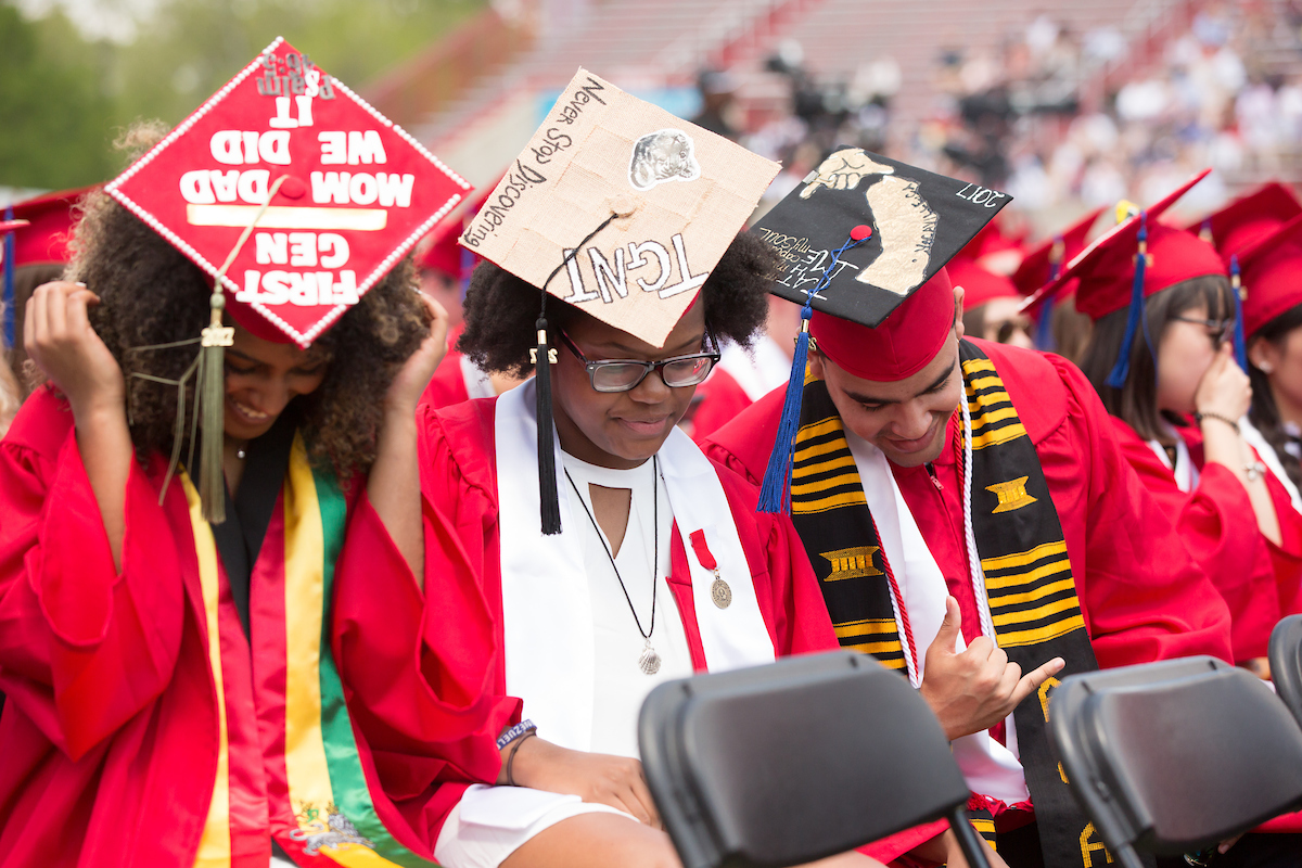 Three students show off their graduation caps at a commencement ceremony, with one reading "First Gen, Wow Dad We Made It."