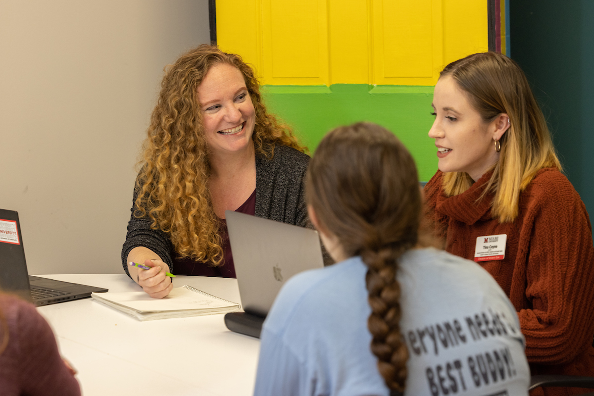 An advisor speaks with two students at a table with a Pride flag in the background.