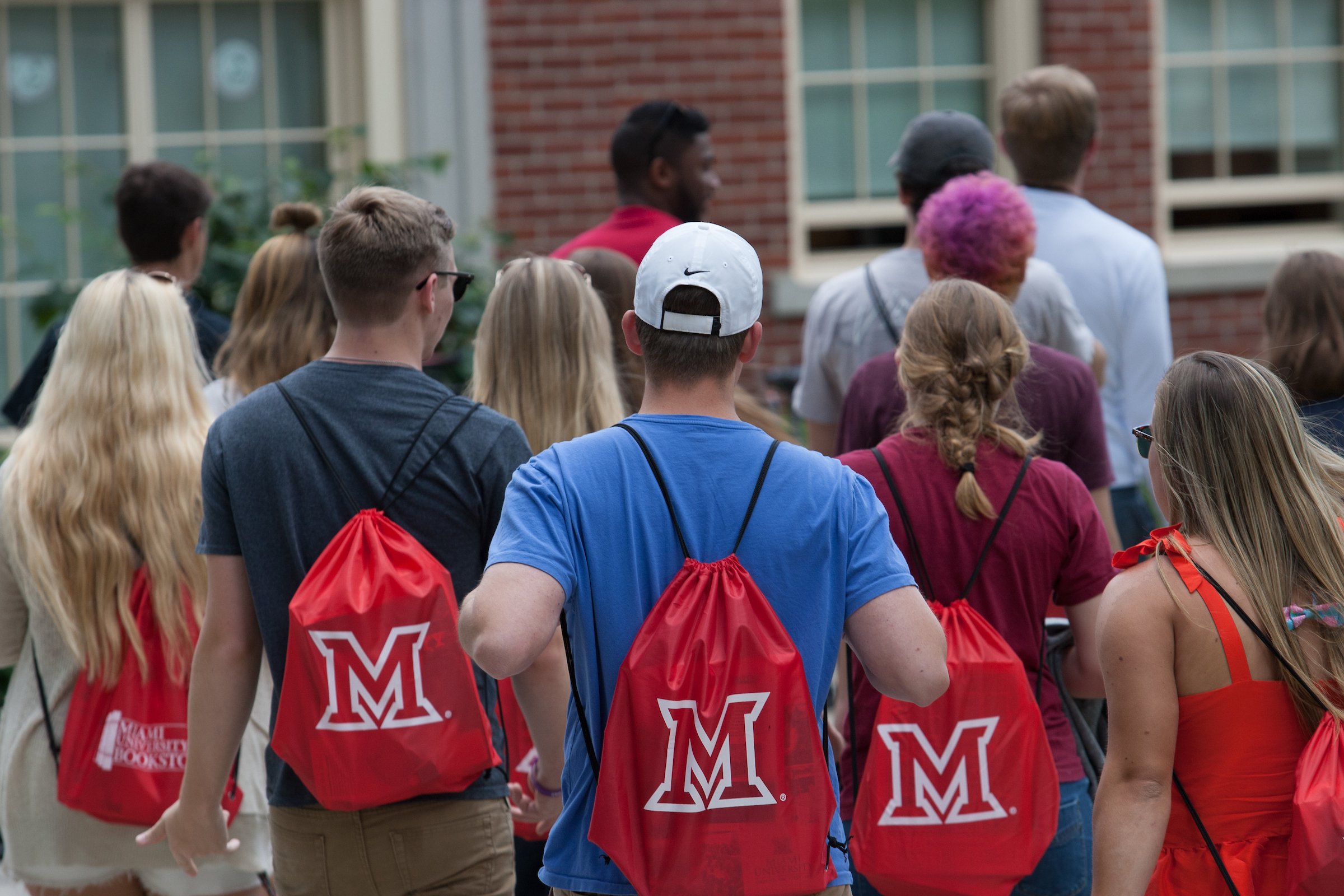 Five students wearing drawstring bags with Miami's logo on their backs stand facing away from the camera outside of a campus building.