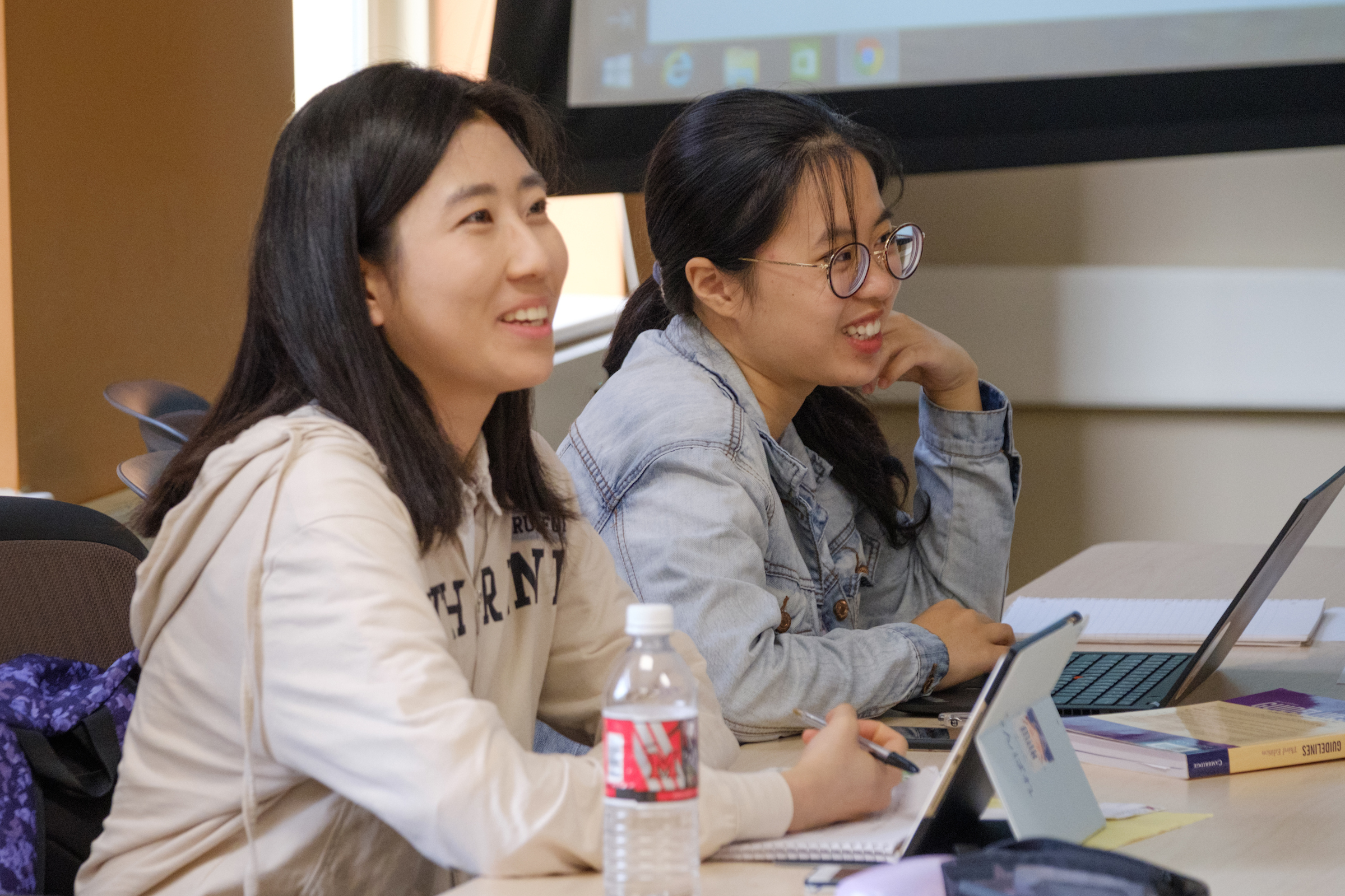 Two students smile while taking notes in a classroom.