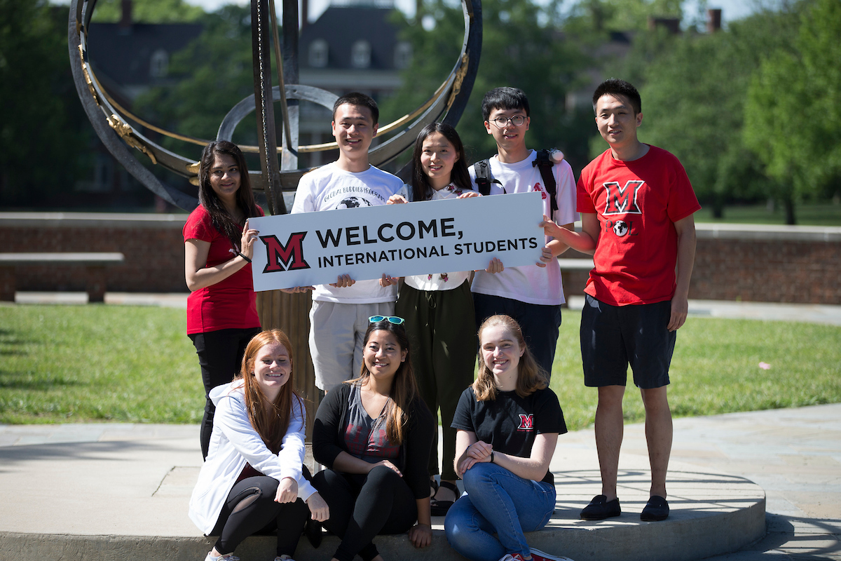 Eight international students pose for a photograph in front of the Tri-Delta Sundial with a sign reading "Welcome International Students."