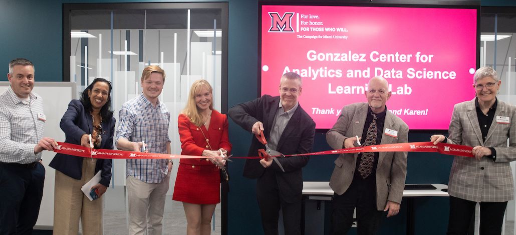 Six people holding and cutting a red ribbon while smiling at the camera.