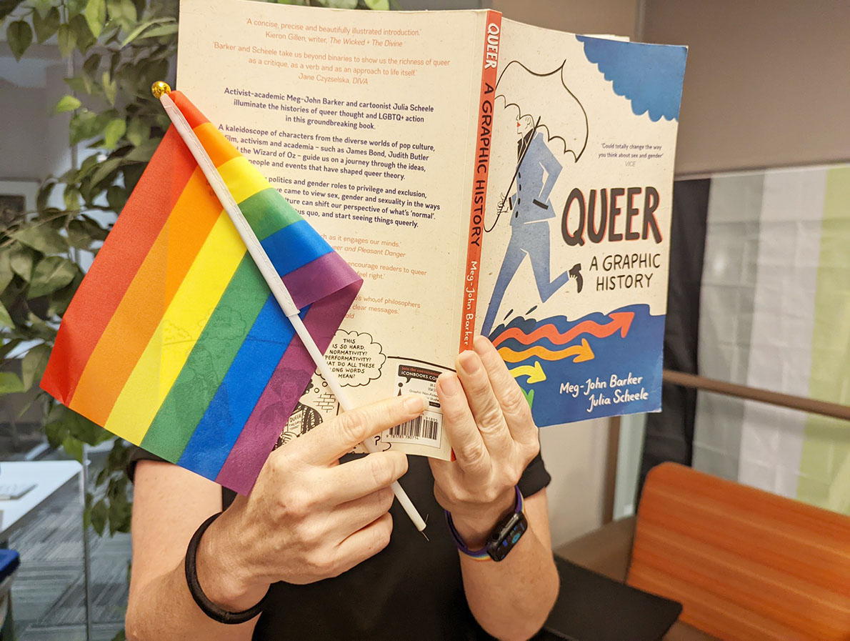 A person holding a queer book and a rainbow flag.