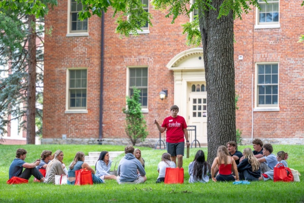A student in a red shirt standing, with a group of about 15 students sitting on the grass around him.