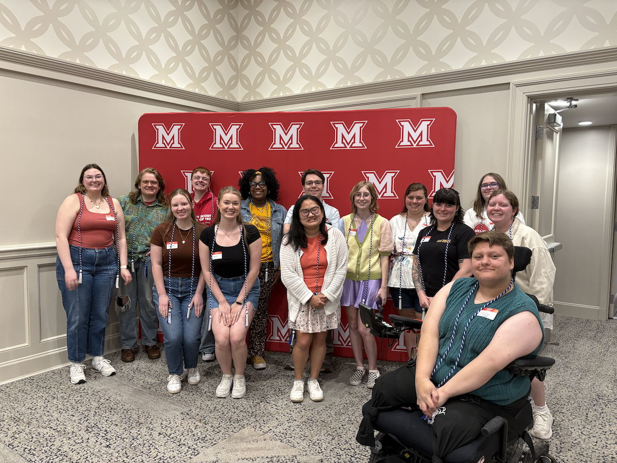 Group of about 12 students, including one in a wheelchair, posing in front of a Miami M background. 