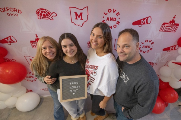 Family of four posing in front of a Miami background. One holds a sign that reads "Proud RedHawk family."