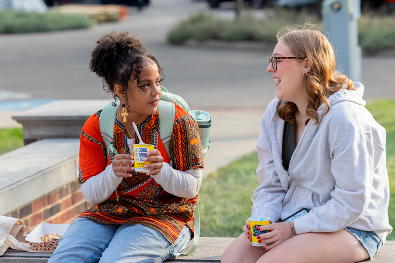 Two students sit outdoors on a low wall, chatting while holding drinks.