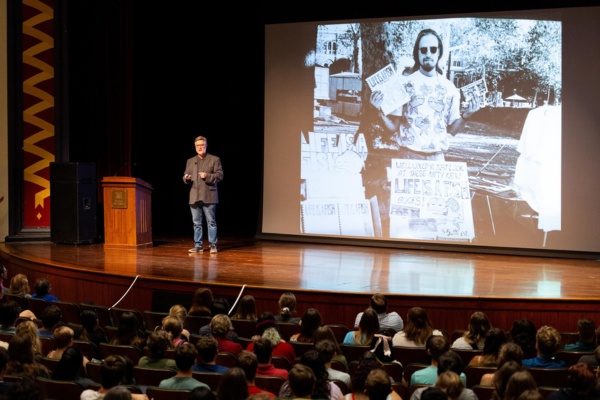 A man on stage in Hall Auditorium, a large screen behind him and a large crowd in the audience.