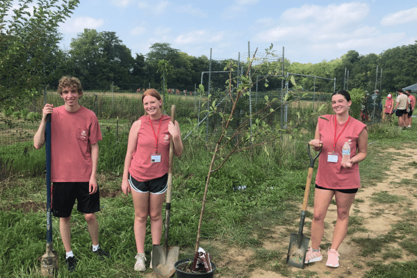 Three female students in red shirts, each holding shovels in a grassy area, preparing for service.