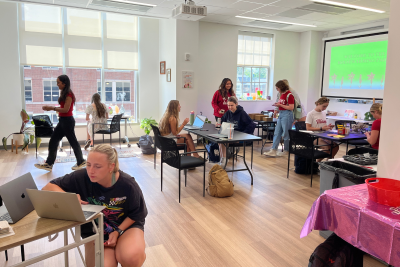 Students working at tables with laptops and craft supplies in a bright classroom with large windows and a projector screen.