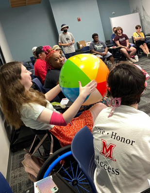 about 20 students in a large circle at desks, one student in a wheelchair is holding a beach ball with questions. 