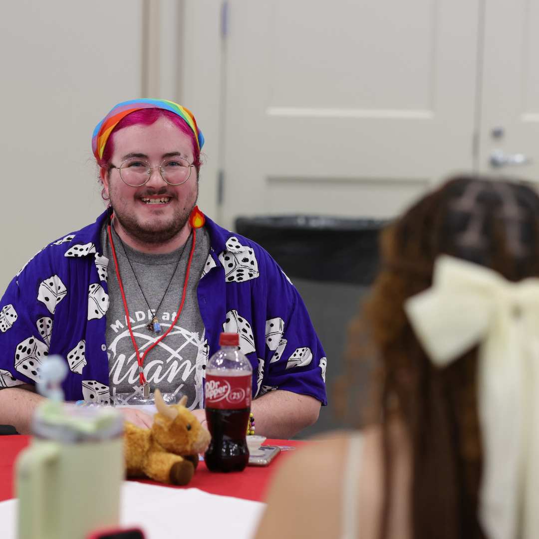 Student wearing a Made at Miami shirt, wearing a colorful rainbow bandana, pink hair and fire earrings. Student is sitting at a table during MADE.