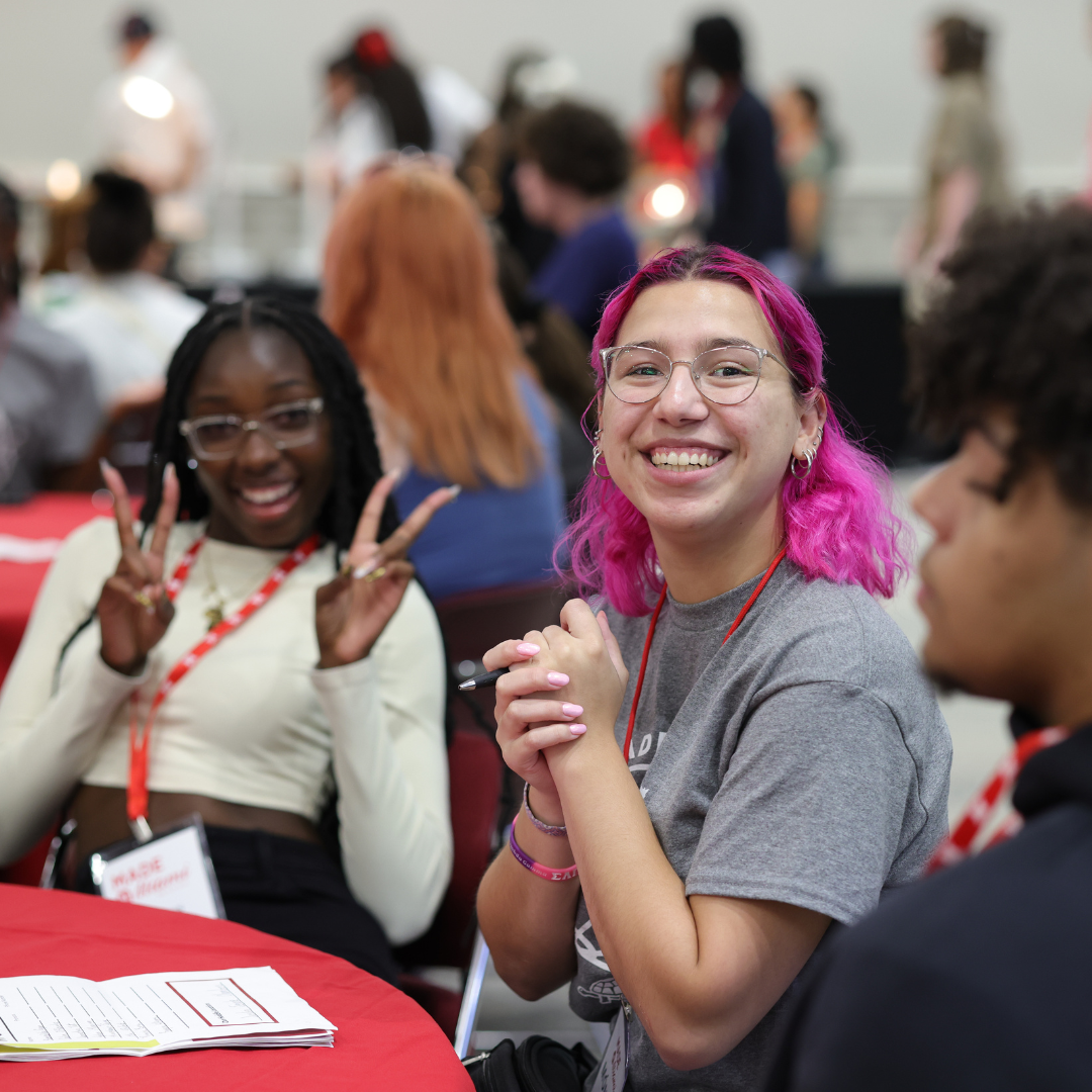 Two students smiling and posing to the camera, sitting at a table during MADE.