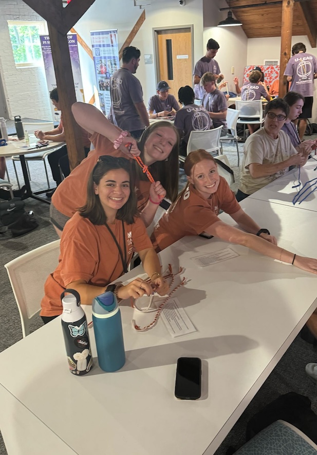 Three students in orange shirts at a table in a cabin-like setting, with many students in different color shirts at different tables behind them. 