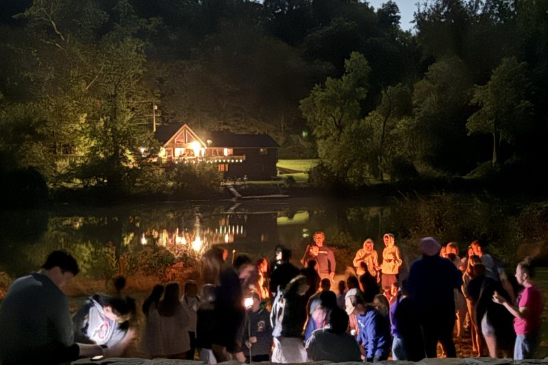 Large group of students huddled around a bonfire on a dark night, lake and cabin in the background.
