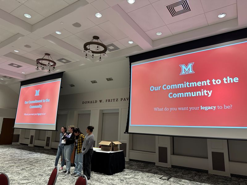 Four students in front of a large group in an Armstrong pavilion room, large screens behind them. 