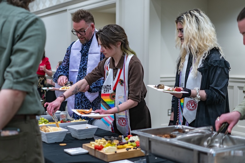 Students and staff in graduation stoles with rainbow flags getting food through a buffet line. .