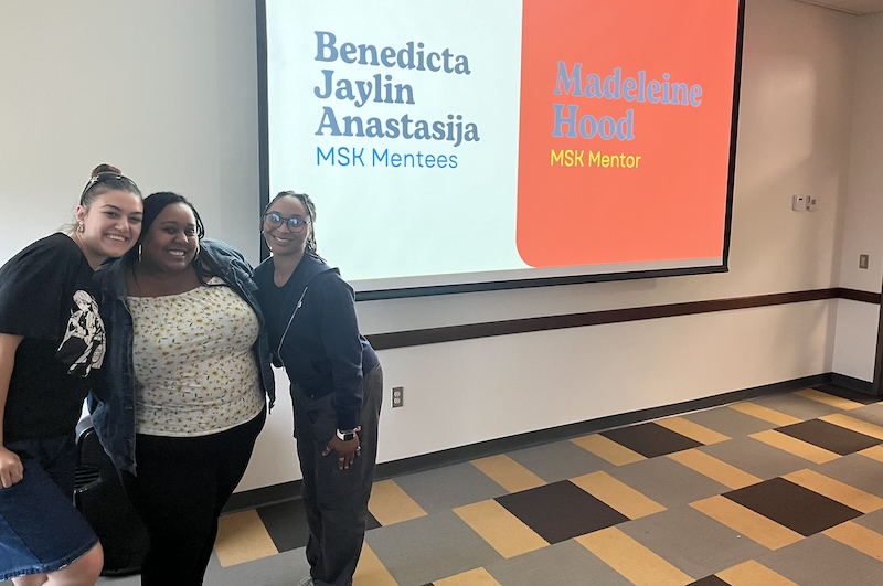 Three women posing in front of a projector screen that reads a mentor's name with the mentee names. 