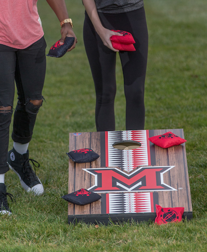 A cornhole set with a large Miami beveled M and a vertical stripe filled with the ribbonwork pattern. Red and black cornhole bags with the Myaamia Heritage Turtle mark on them