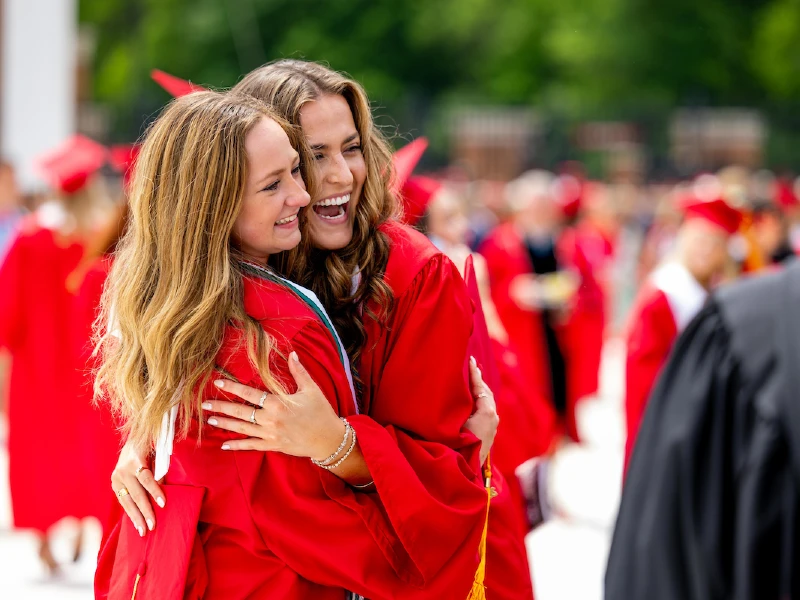 Two female students at graduation wearing red gowns and hugging.