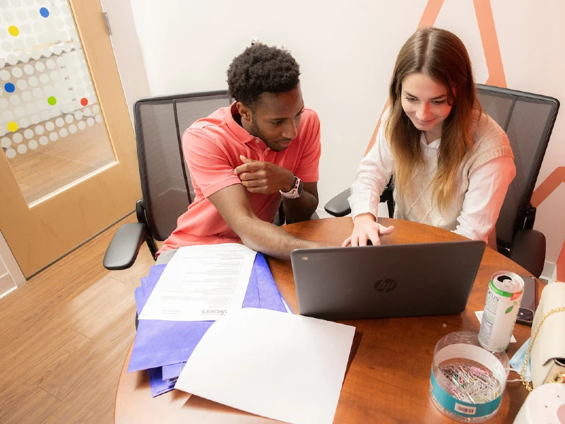 Two students working in office looking at a laptop on desk with purple folders and paper.