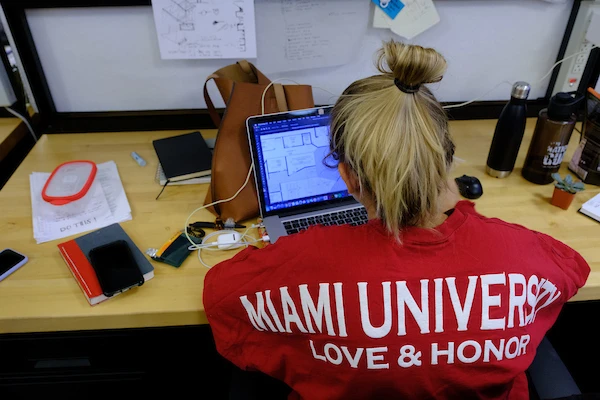 Student working at desk with back to camera wearing Miami University shirt.