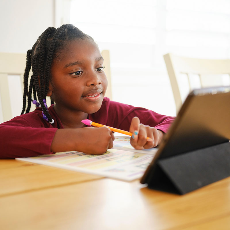 A young student completes a workbook while using a tablet in the classroom.