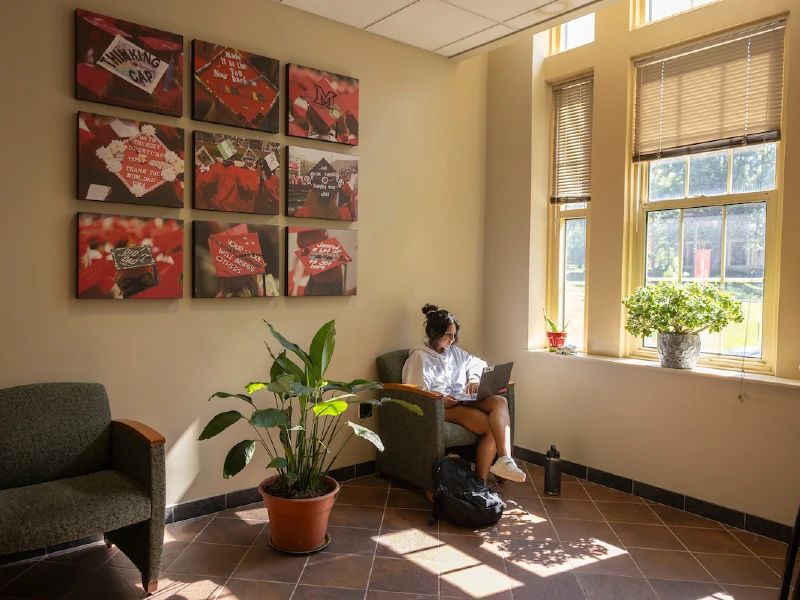 A student holding laptop in lobby, with pictures of graduation caps hanging on wall.