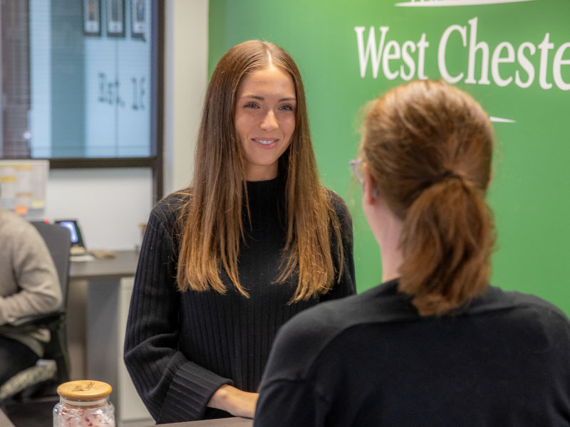 Student working in an office checking someone in at a front desk.
